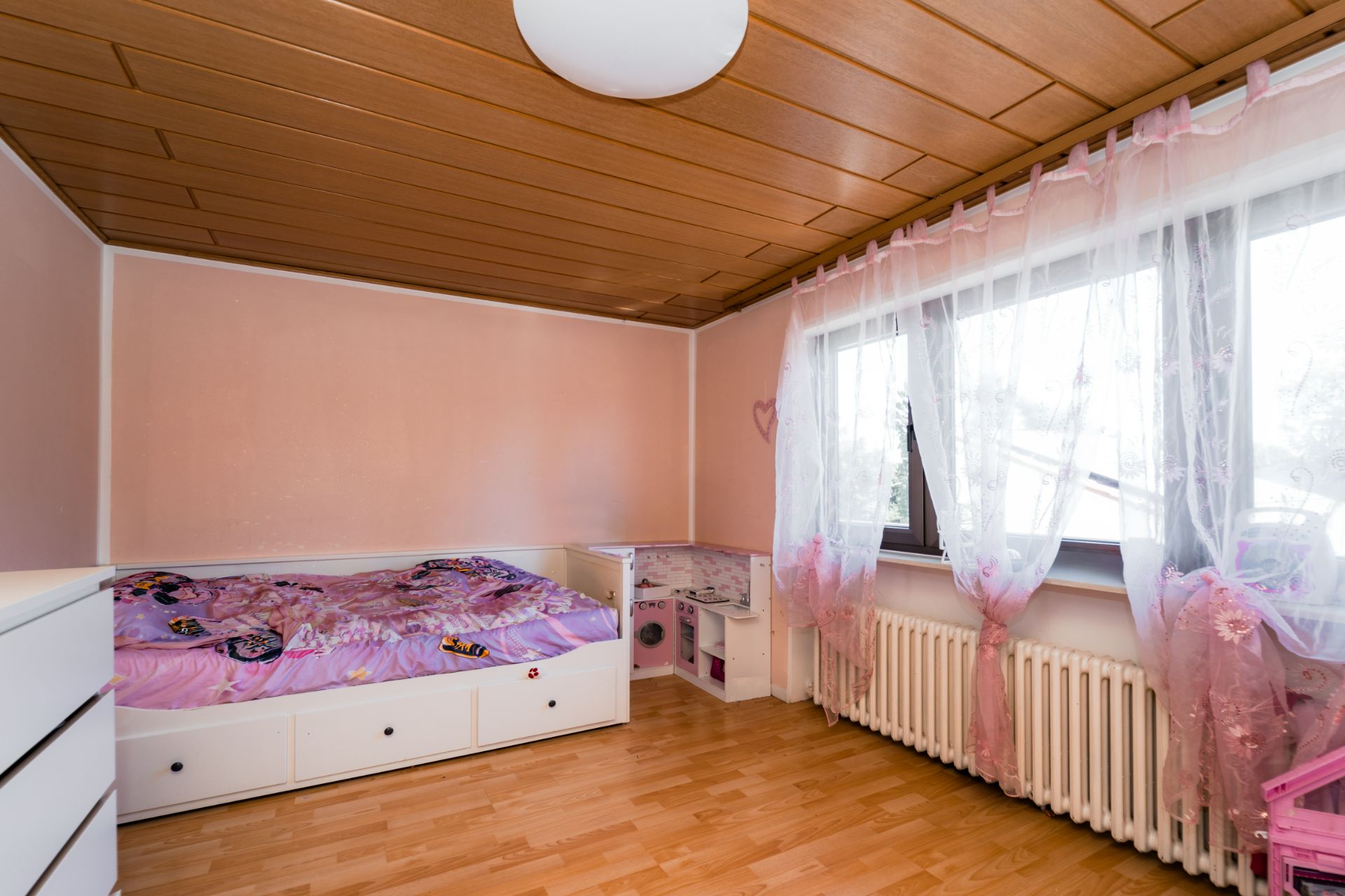 Helles Kinderzimmer mit rosa Wänden, Einzelbett, Schreibtisch und großen Fenstern mit Vorhängen.
