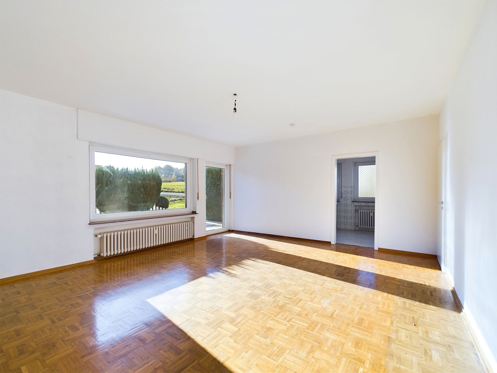 Heller, leerer Raum mit Parkettboden, großem Fenster und Tür zur Terrasse.