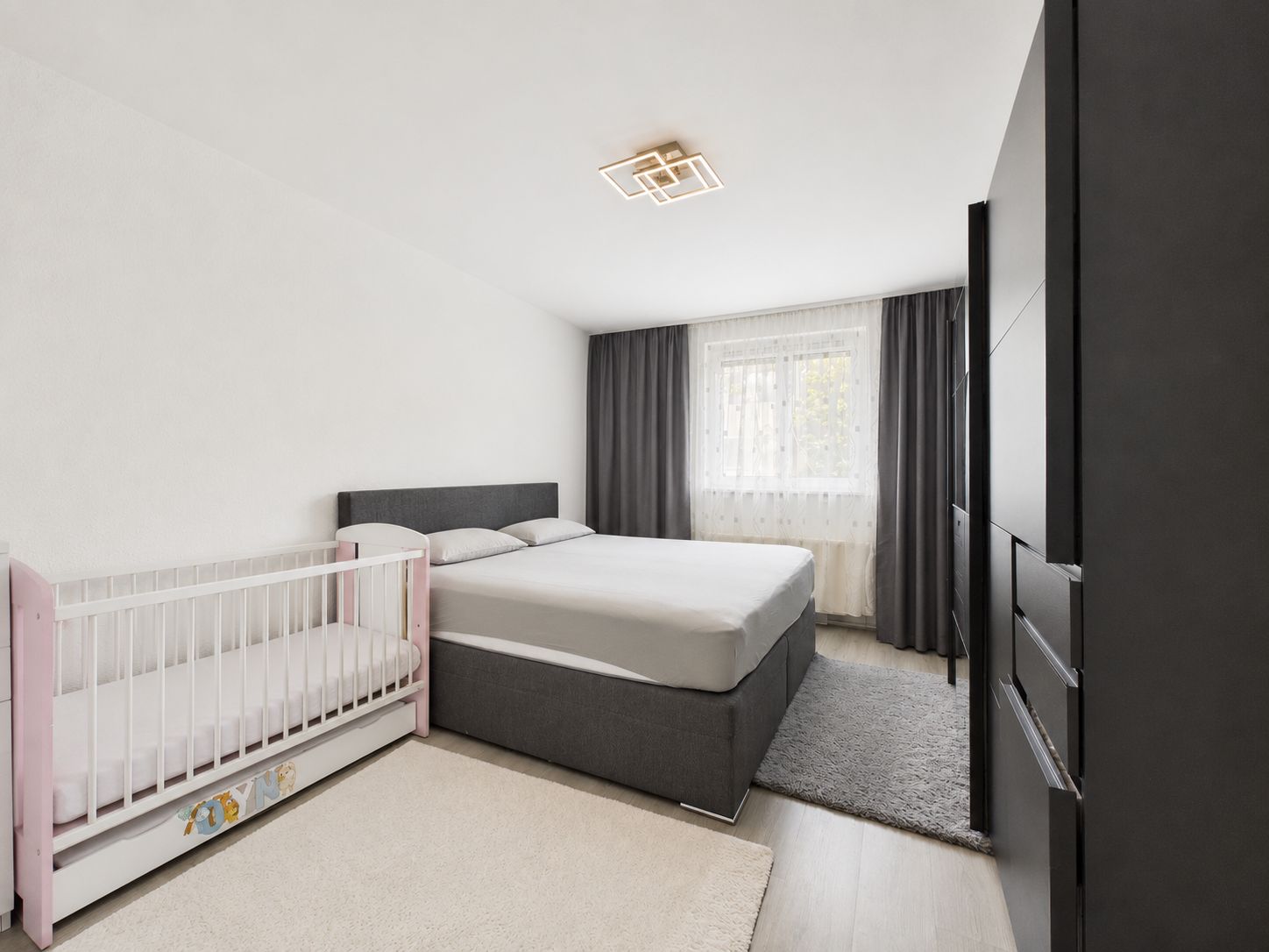 Schlafzimmer mit Doppelbett, Babybett, grauem Schrank und Fenster mit Vorhängen.