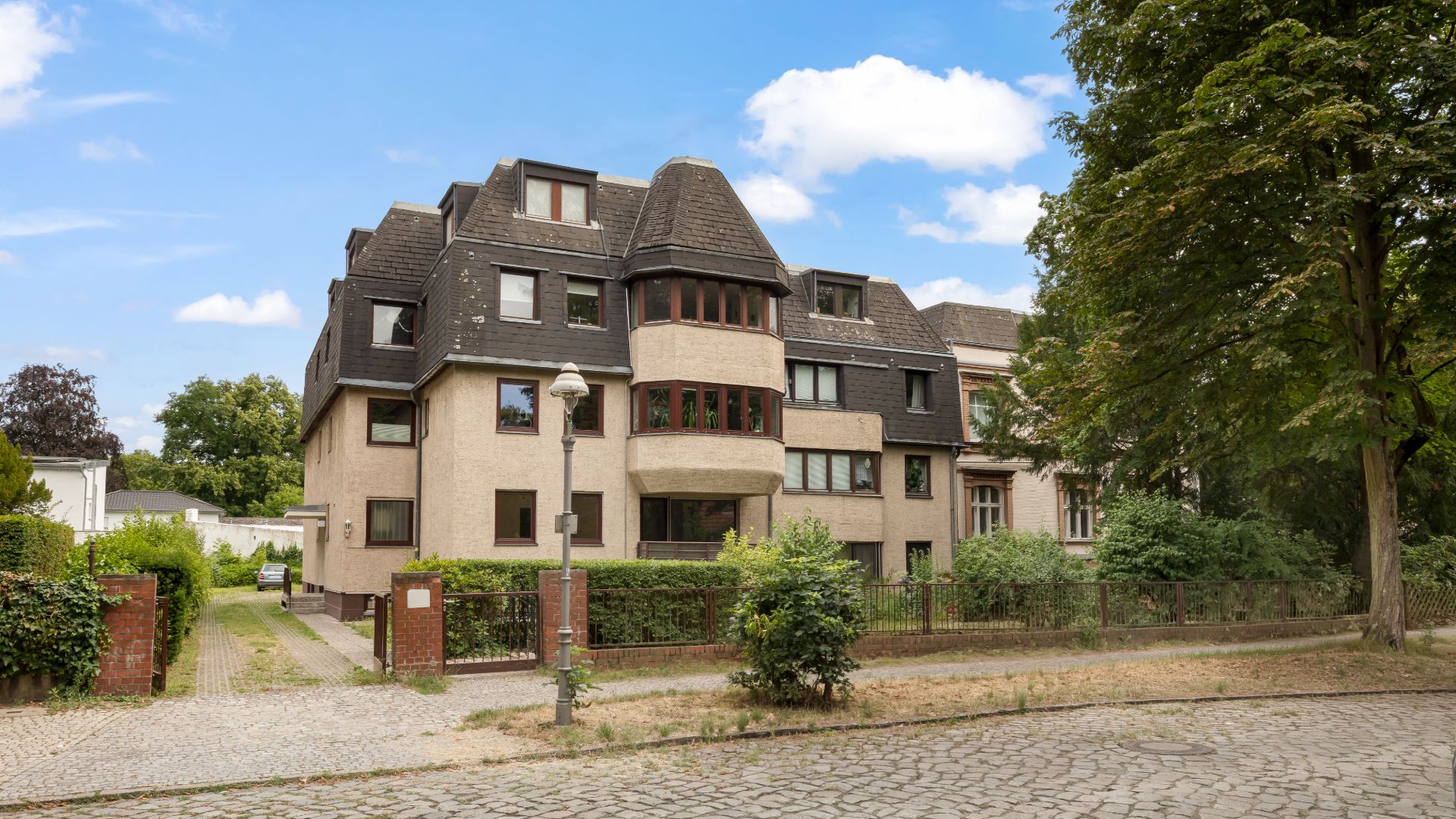 Mehrfamilienhaus mit dunklem Dach und Vorgarten an einer gepflasterten Straße, umgeben von Bäumen.