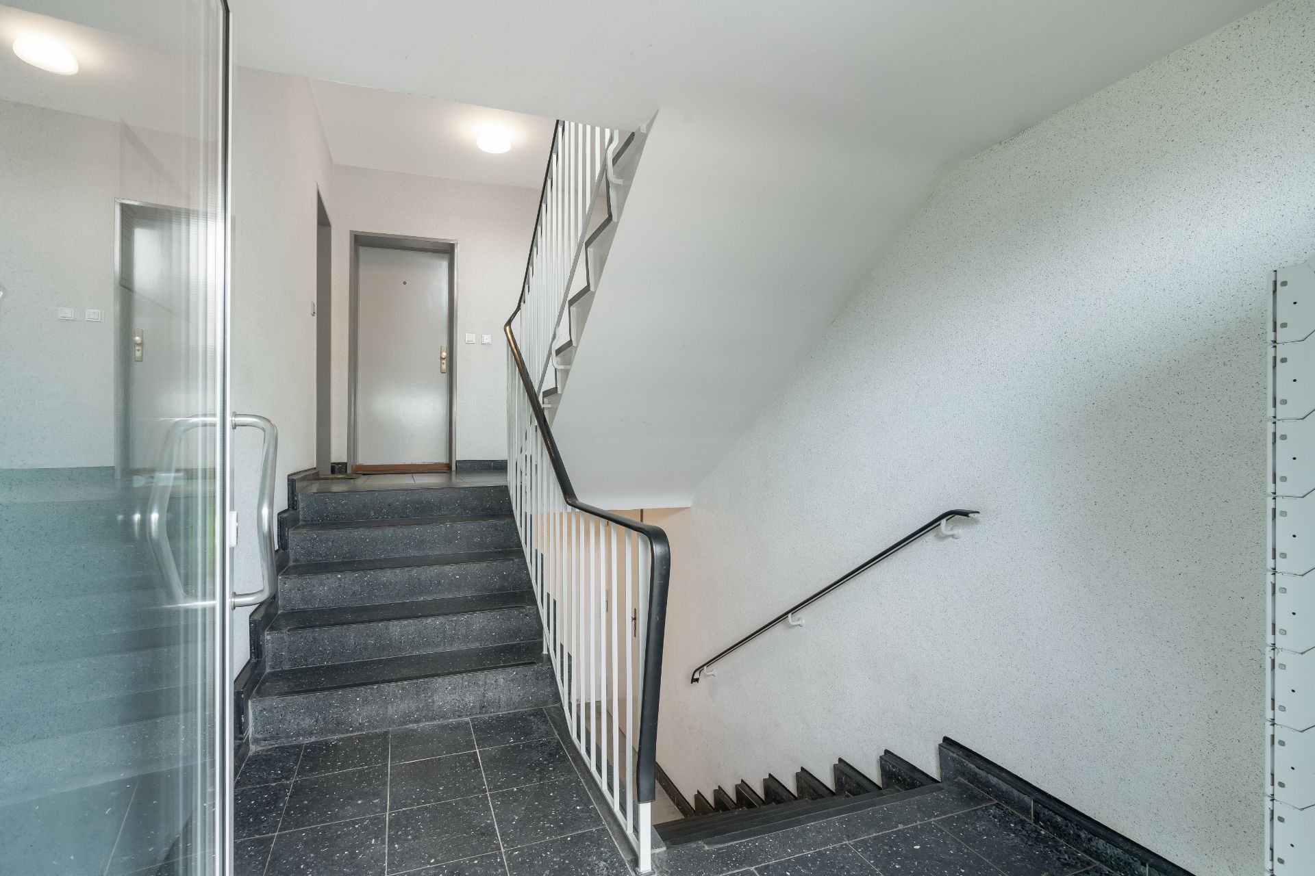 Helles Treppenhaus mit schwarzen Stufen, Handläufen und einer Wohnungstür im Hintergrund.