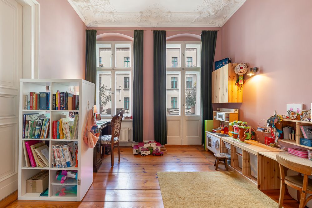 Helles Kinderzimmer mit Bücherregal, Schreibtisch, Spielzeug und großen Fenstern mit dunklen Vorhängen.