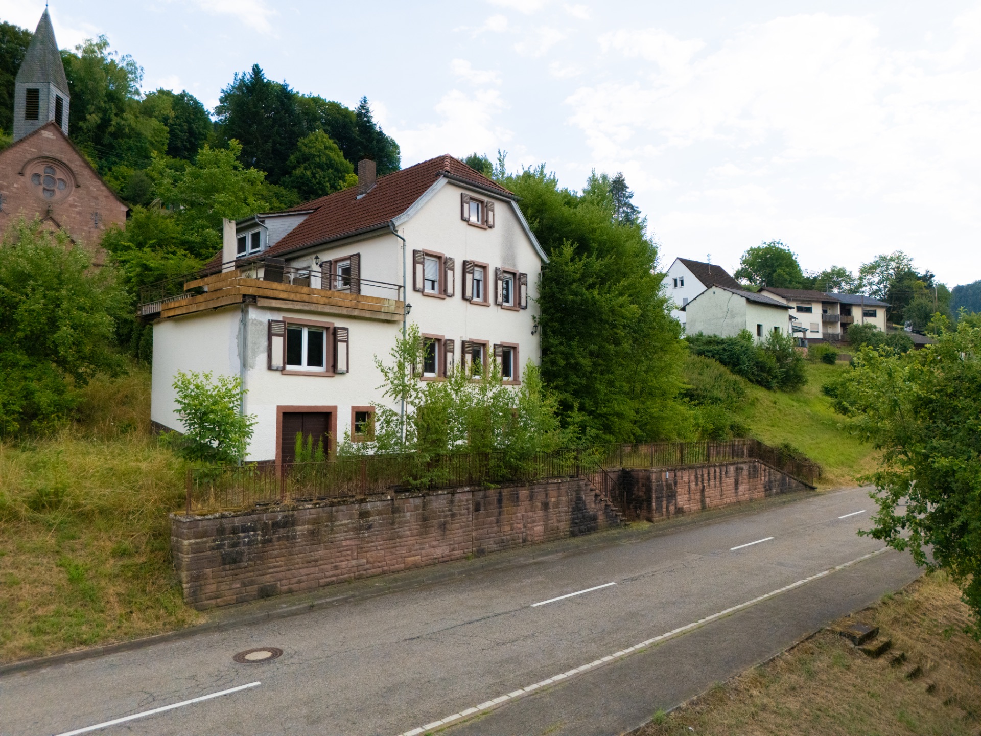 Weißes Wohnhaus mit braunem Dach und Fensterläden an einer Straße, umgeben von Bäumen.
