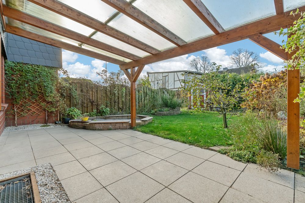 Überdachte Terrasse mit Fliesenboden, Blick auf Garten mit Rasen, Beeten und Holzzaun.