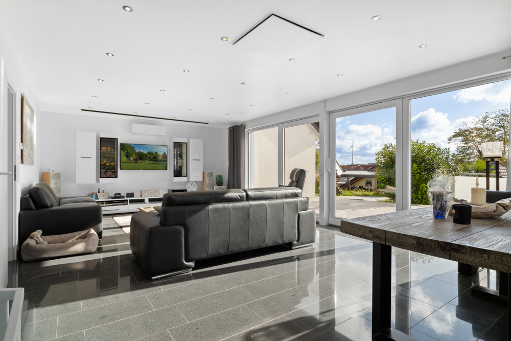 Helles Wohnzimmer mit schwarzen Sofas, großem Fernseher und Glasfront zum Garten.