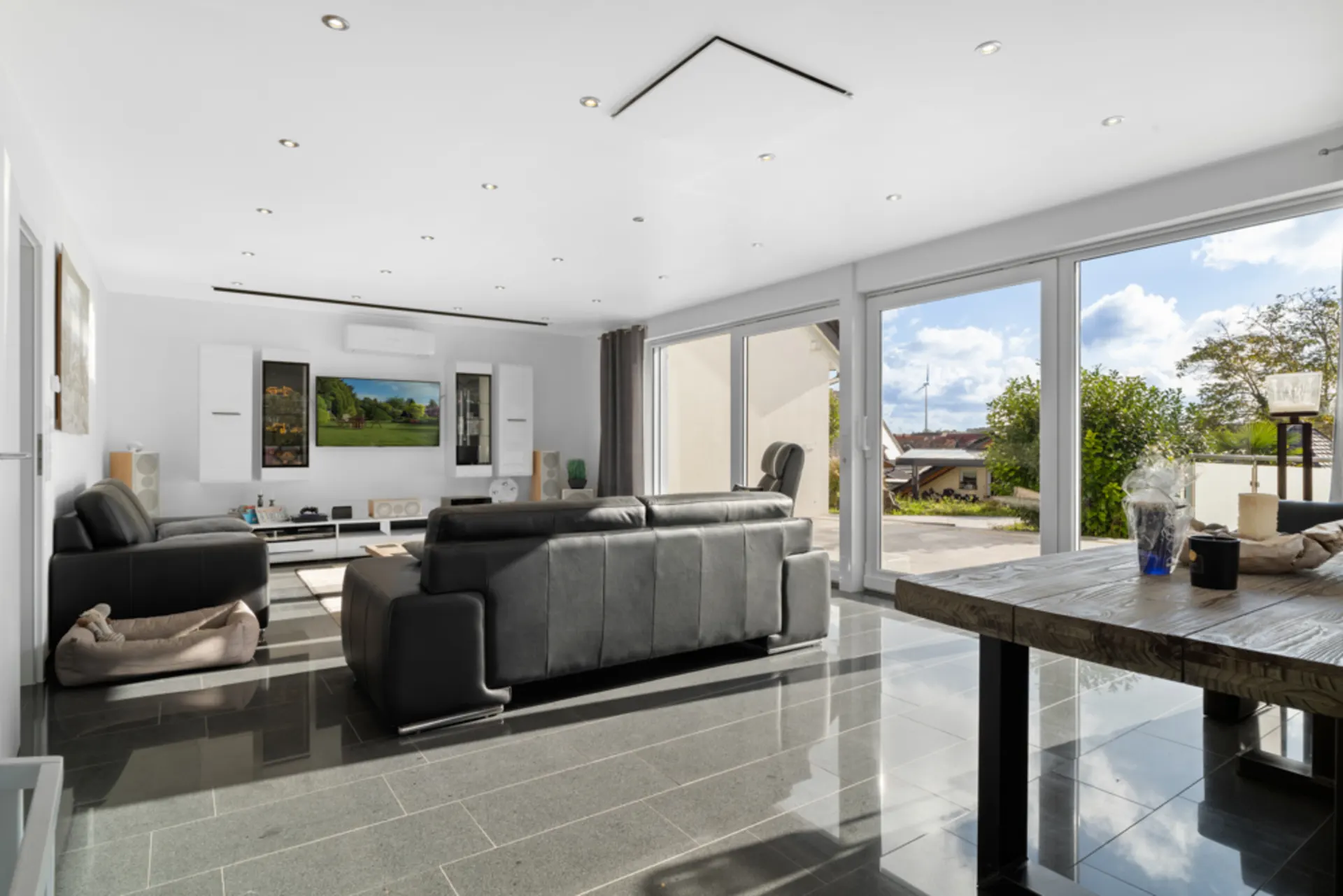 Helles Wohnzimmer mit schwarzen Sofas, großem Fernseher und Glasfront zum Garten.