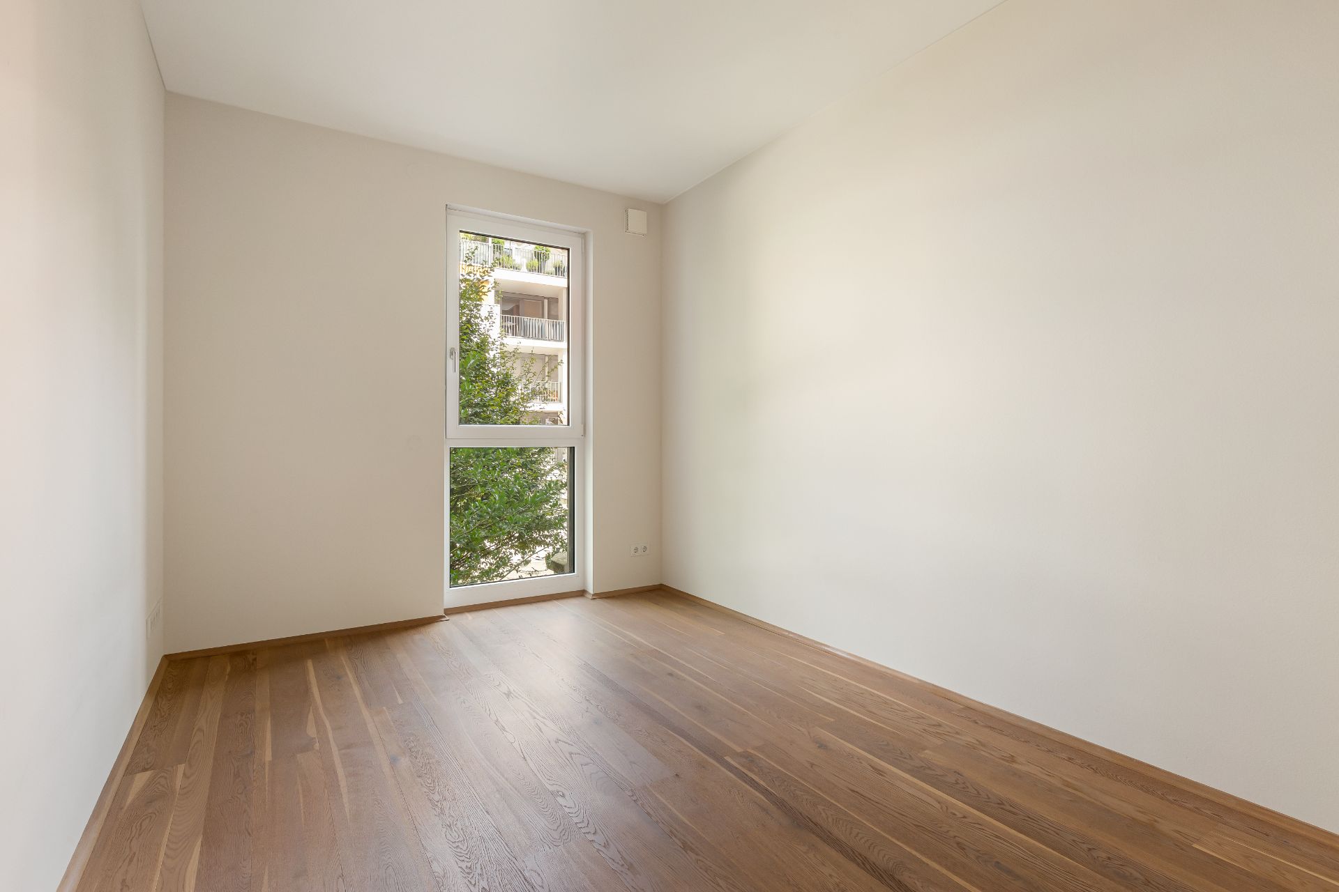 Leerer Raum mit Holzfußboden, weißer Wand und Fenster mit Blick auf Bäume.