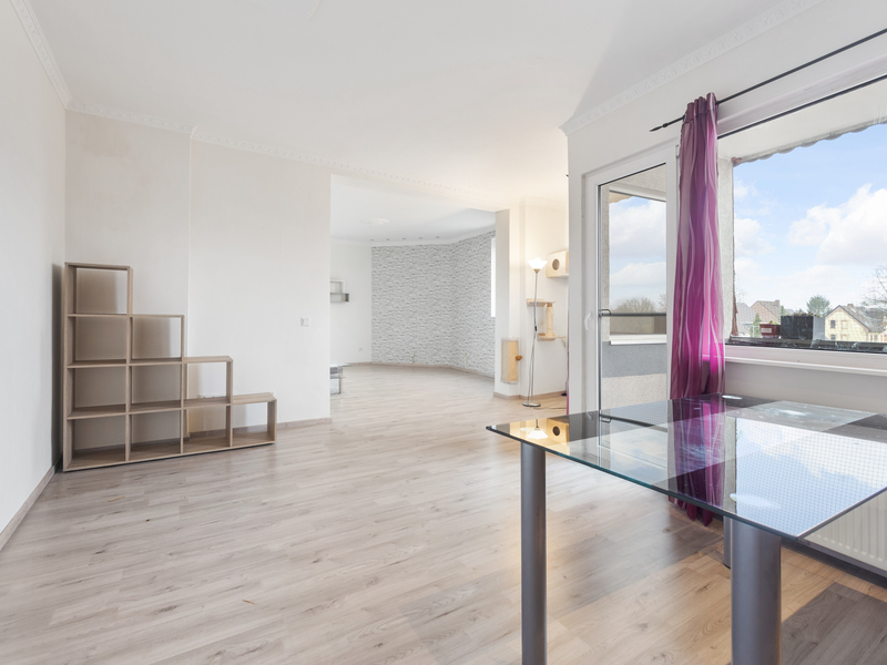 Helles Wohnzimmer mit Glasstisch, Regal, Holzboden und großem Fenster mit lila Vorhang.