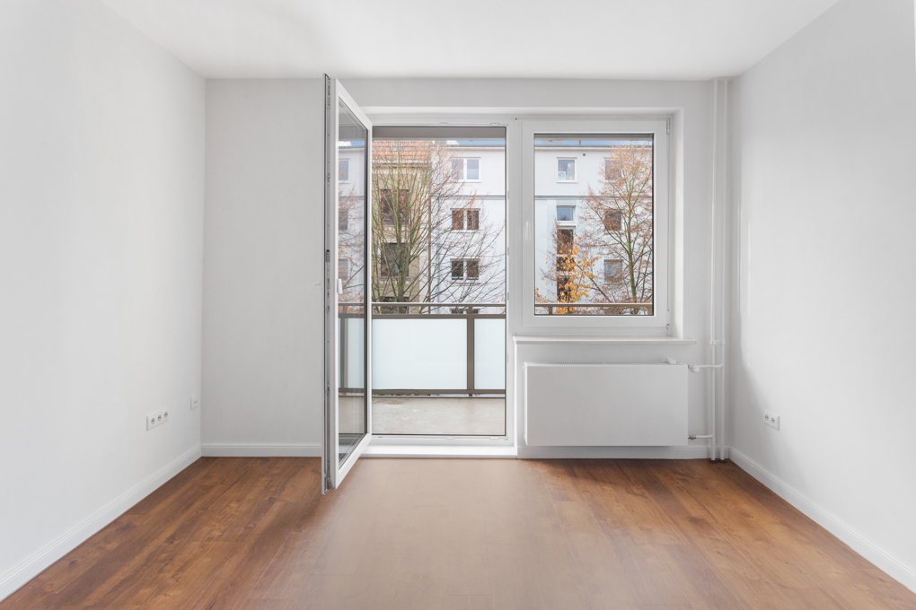 Leeres Zimmer mit Holzboden, weißen Wänden und offener Balkontür.