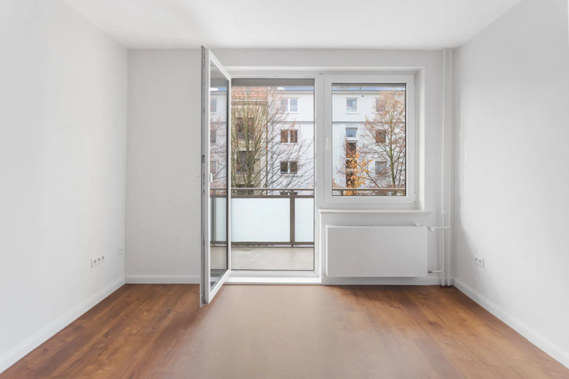 Leeres Zimmer mit Holzboden, weißen Wänden und offener Balkontür.