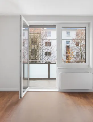 Leeres Zimmer mit Holzboden, weißen Wänden und offener Balkontür.