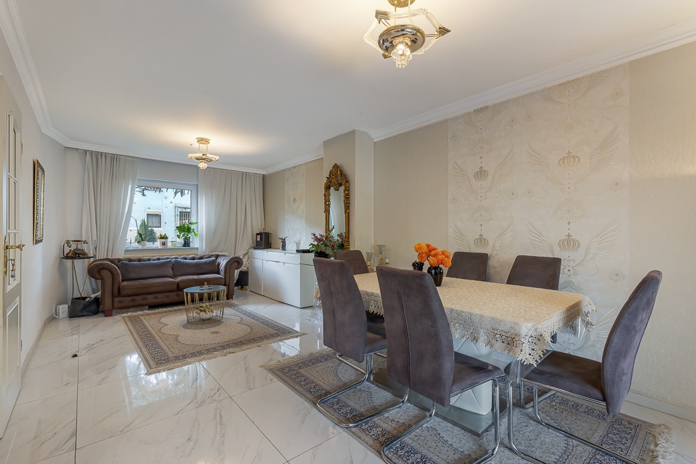 Wohnzimmer mit braunem Sofa, Esstisch mit sechs Stühlen und hellen Fliesenboden.