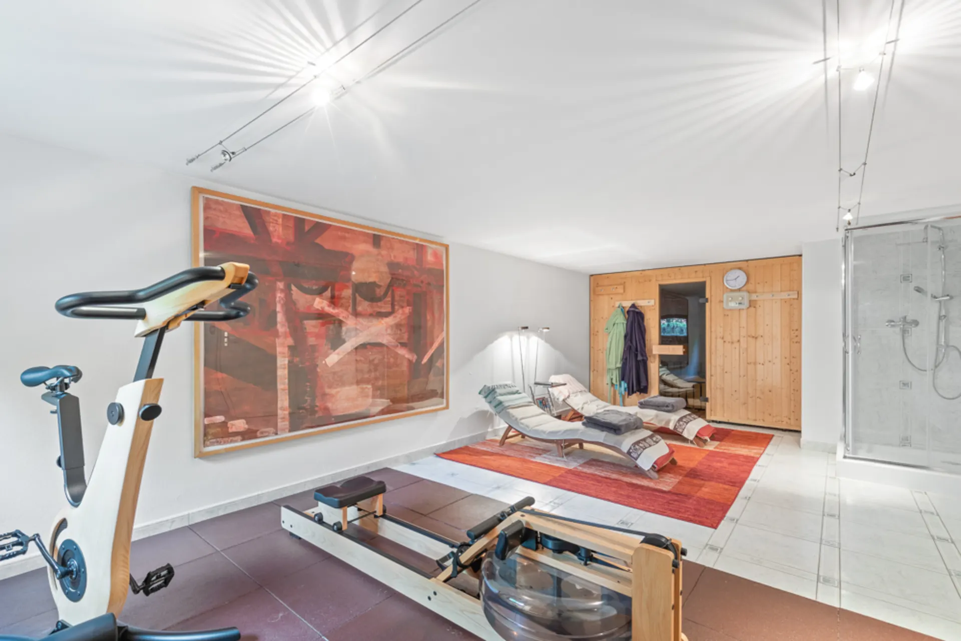 Fitnessbereich mit Ergometer, Rudergerät, zwei Liegen, Sauna und Dusche in einem Raum.