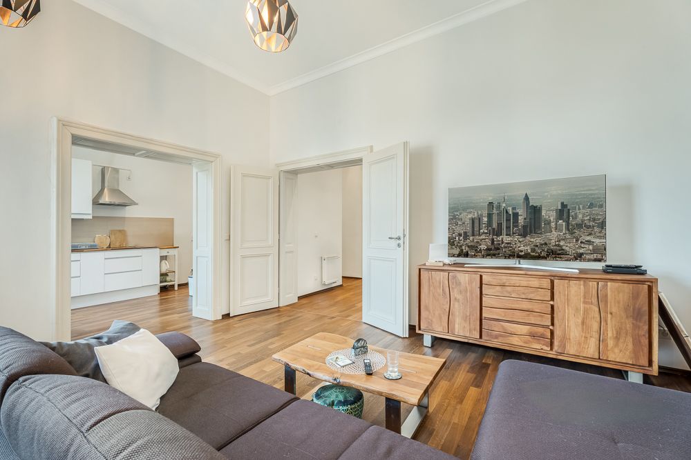Helles Wohnzimmer mit Sofa, Holztisch, Sideboard und Fernseher, angrenzend eine offene Küche.