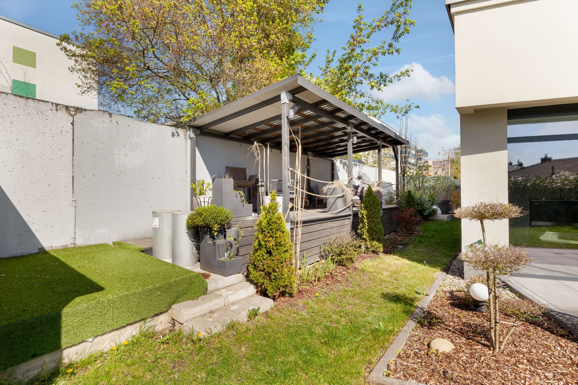 Überdachte Terrasse mit Sitzmöbeln im Garten, umgeben von Pflanzen und Bäumen.