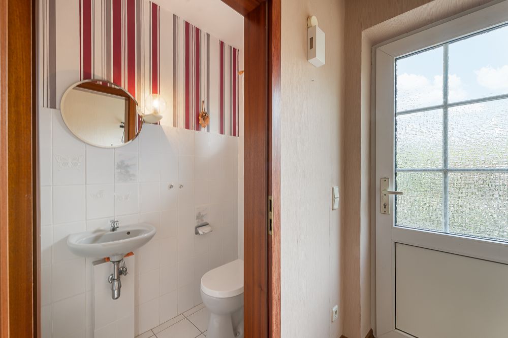Kleines Bad mit WC, Waschbecken, rundem Spiegel und gestreifter Tapete, neben einer Glastür.