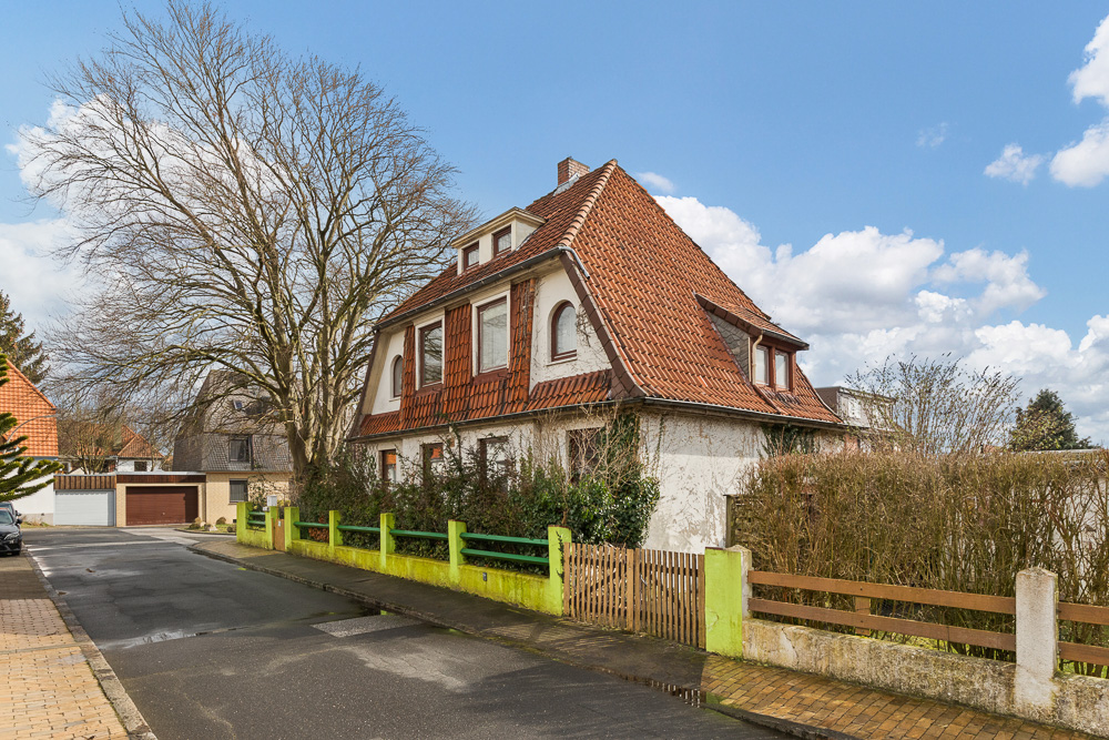 Zweistöckiges Haus mit rotem Dach, grünem Zaun und großem Baum an einer ruhigen Straße.