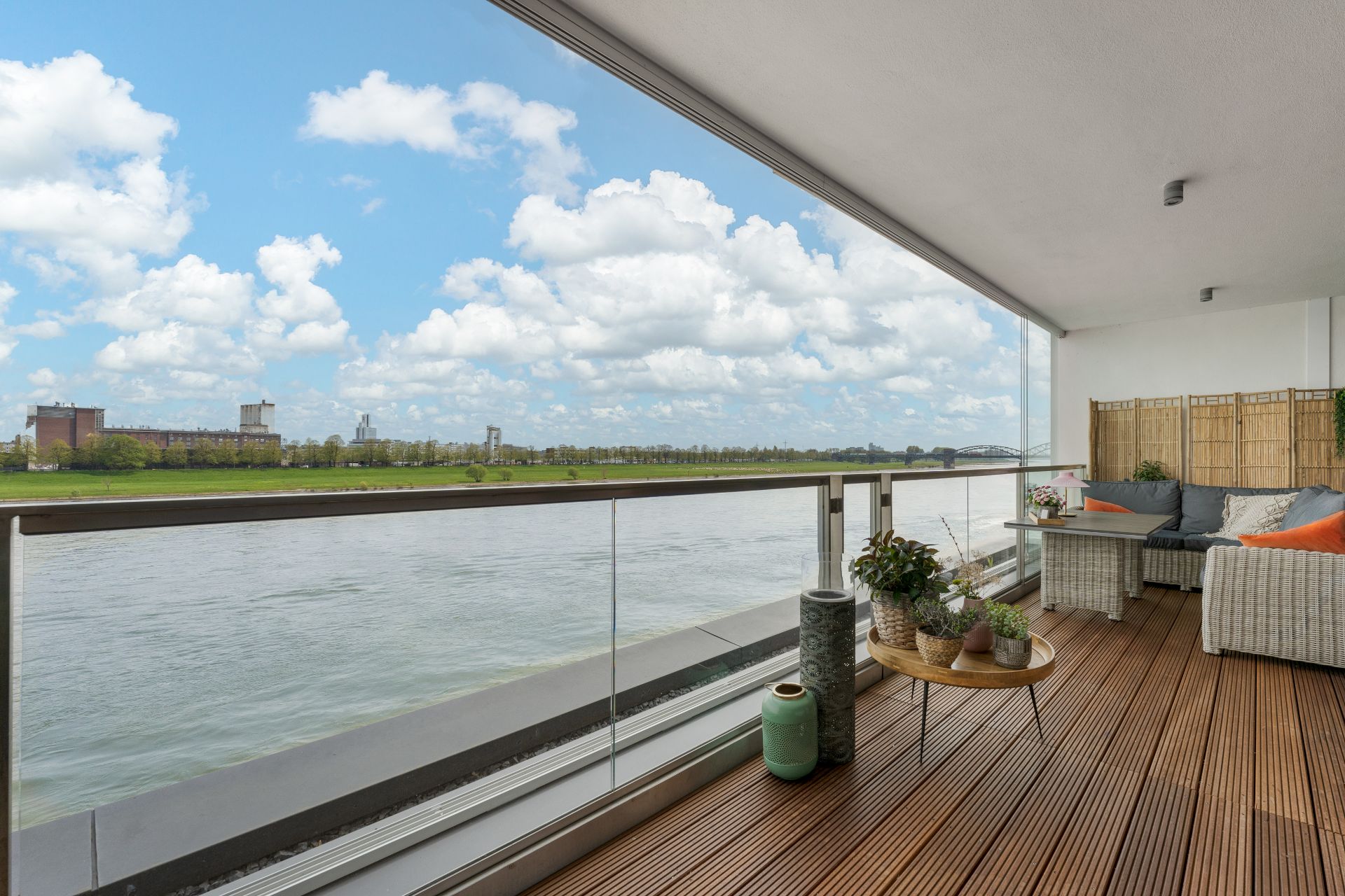 Balkon mit Holzboden, Sitzgruppe und Pflanzen, Blick auf Fluss und grüne Uferlandschaft.