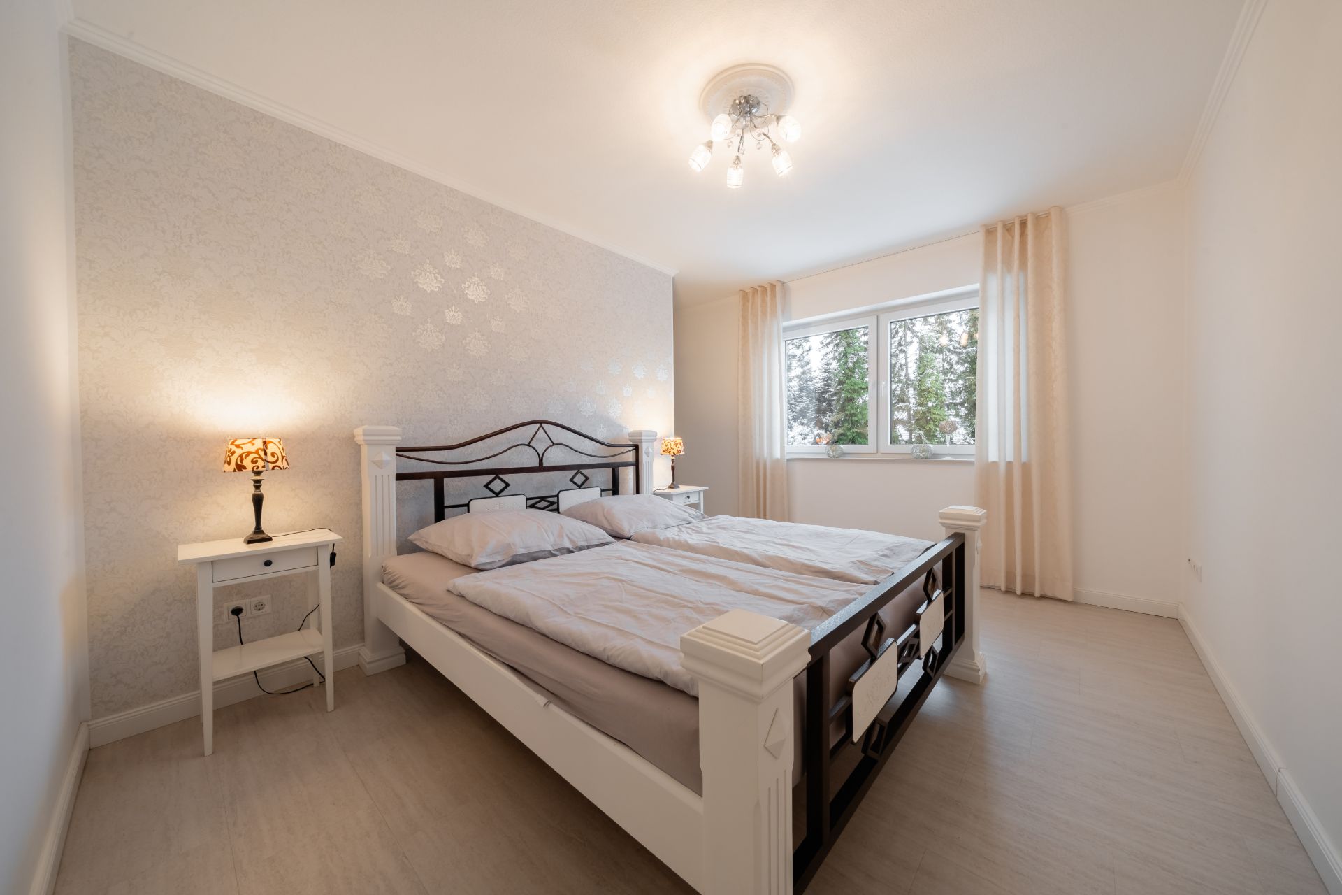 Helles Schlafzimmer mit Doppelbett, zwei Nachttischen und Fenstern mit Vorhängen.