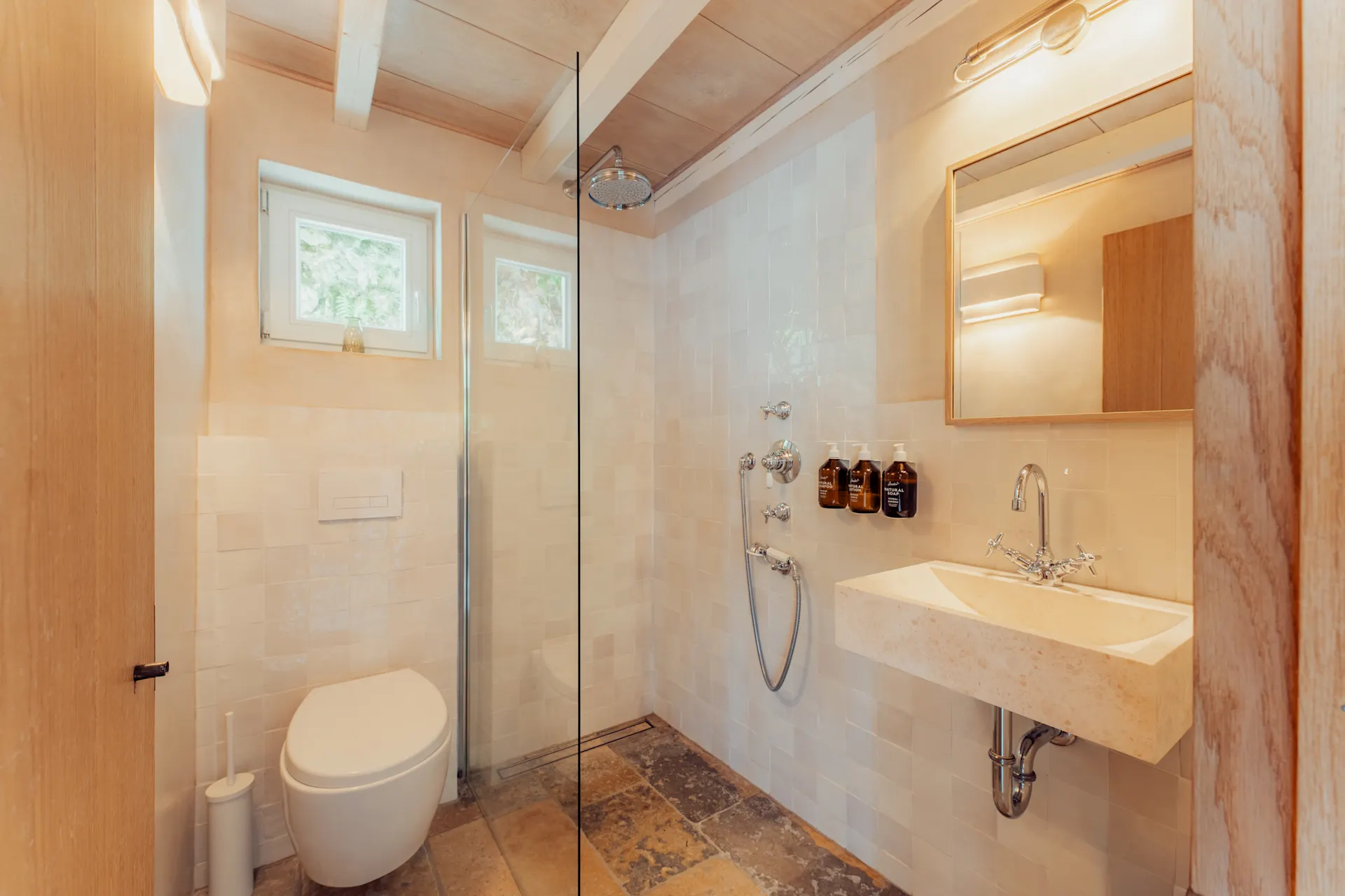 Helles Badezimmer mit Toilette, Dusche, Waschbecken und Spiegel, Fliesenboden und zwei kleinen Fenstern.