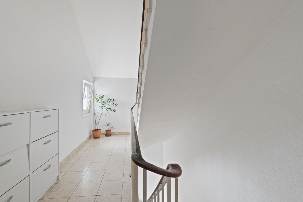 Heller Flur mit Treppengeländer, weißen Schränken und Topfpflanzen am Fenster.