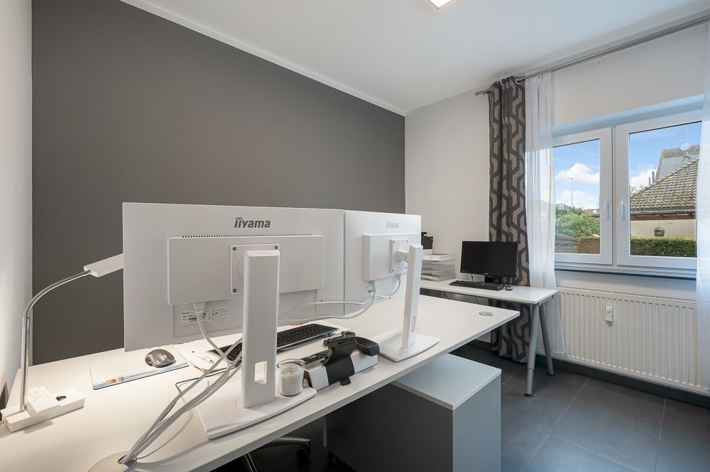 Modernes Arbeitszimmer mit zwei Monitoren, Schreibtischen und großem Fenster mit Tageslicht.