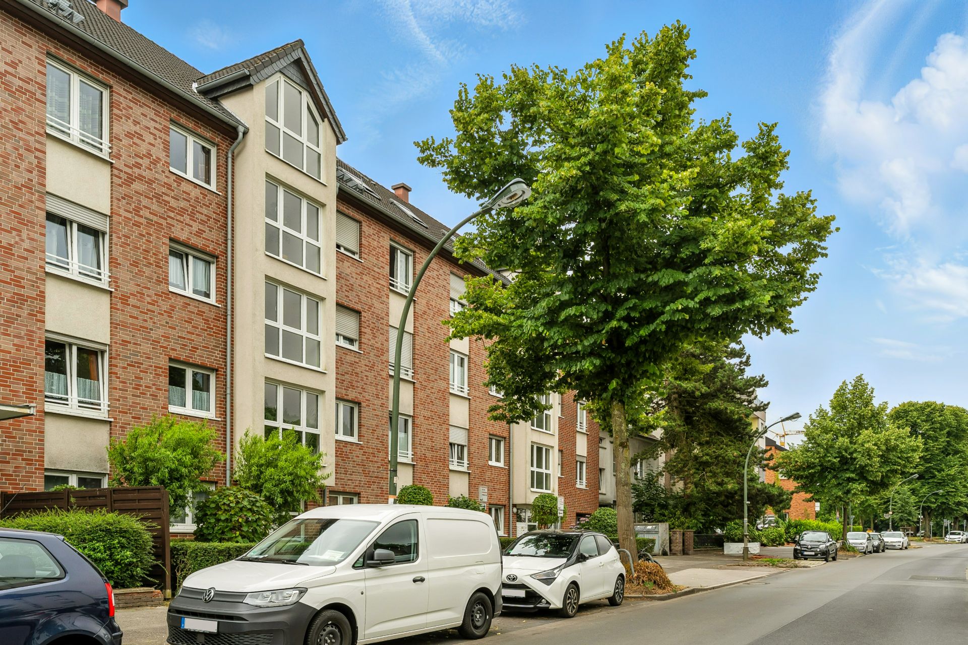 Mehrfamilienhaus mit roten Ziegeln, Bäumen und parkenden Autos an der Straße.