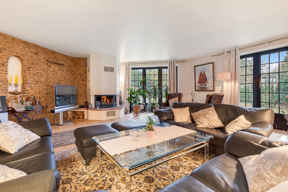 Helles Wohnzimmer mit Leder-Sofas, Teppich, Kamin und großen Fenstern mit Blick ins Grüne.