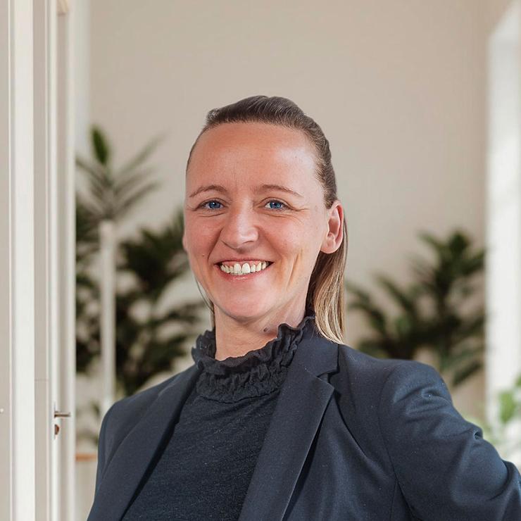 Frau mit dunklem Blazer steht in einem hellen Raum mit Pflanzen im Hintergrund.