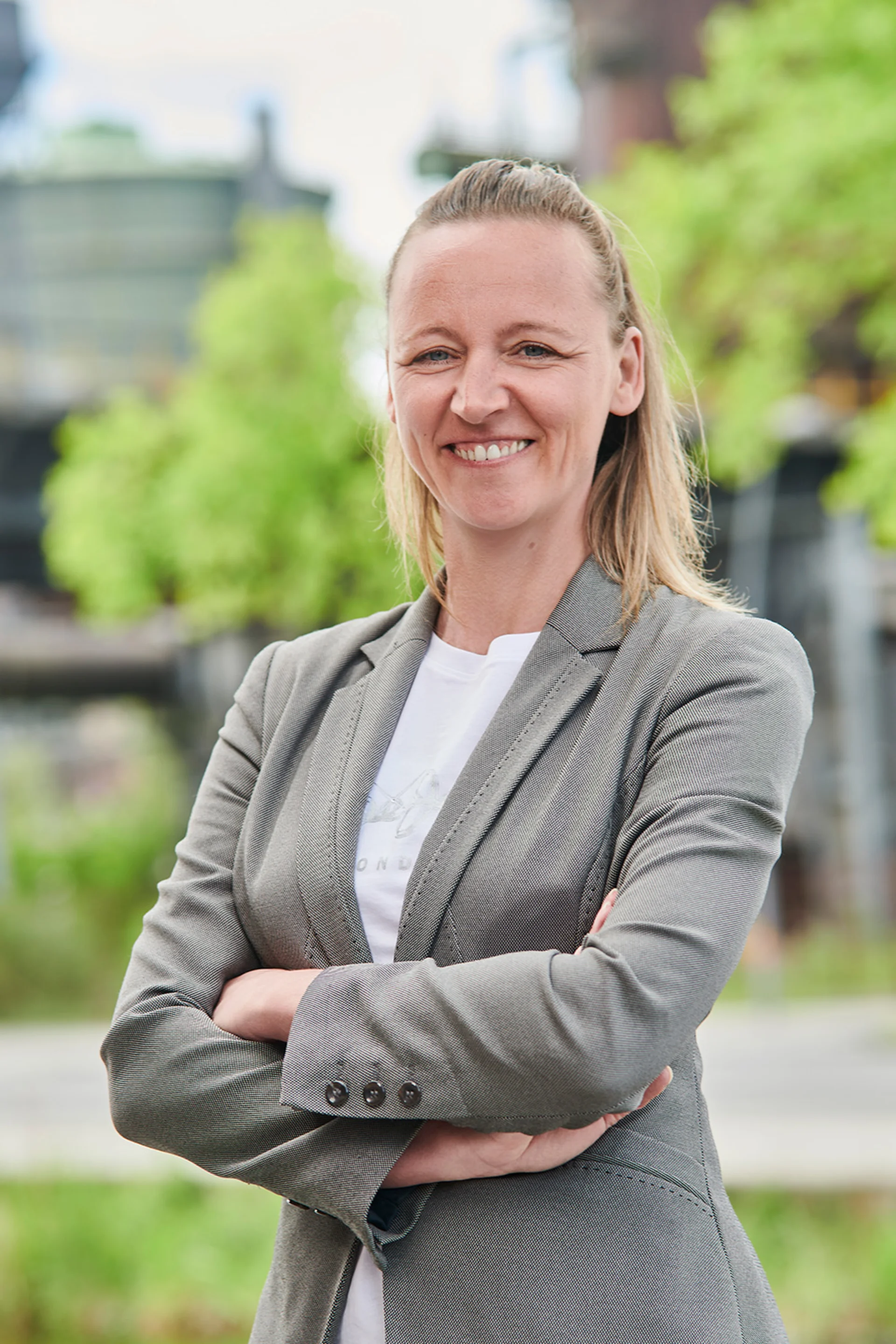 Frau mit dunklem Blazer steht in einem hellen Raum mit Pflanzen im Hintergrund.