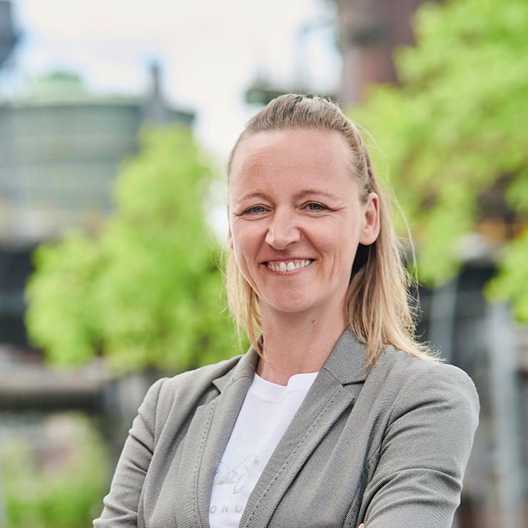 Frau mit dunklem Blazer steht in einem hellen Raum mit Pflanzen im Hintergrund.