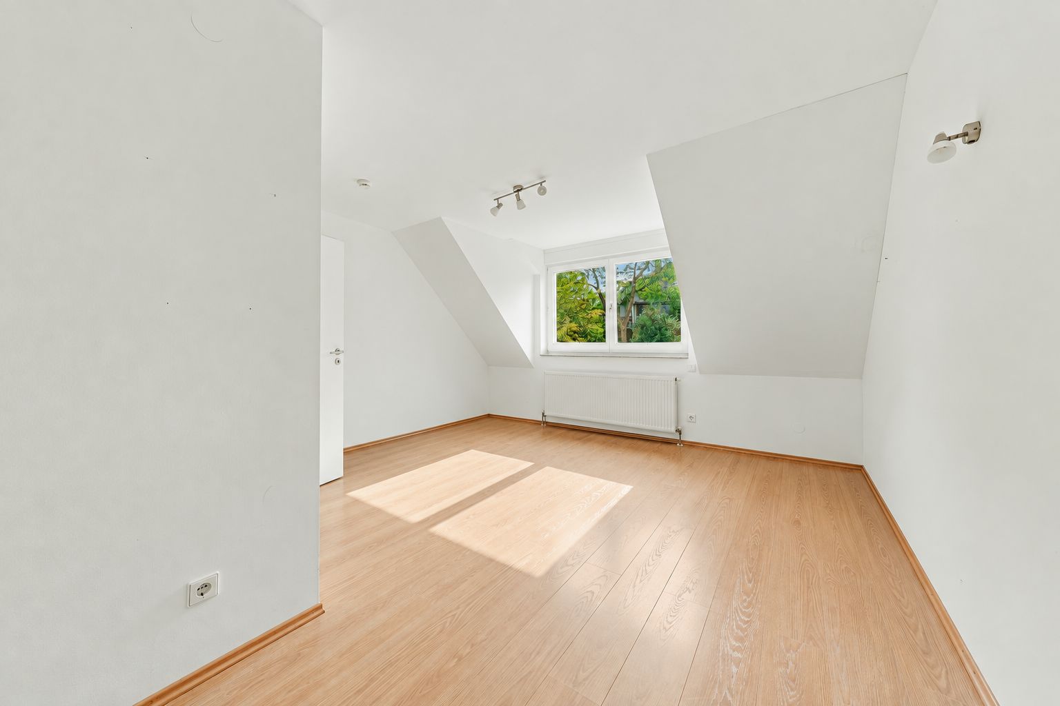 Helles Zimmer mit Dachschräge, weiß gestrichenen Wänden und Holzfußboden, Fenster mit Blick ins Grüne.