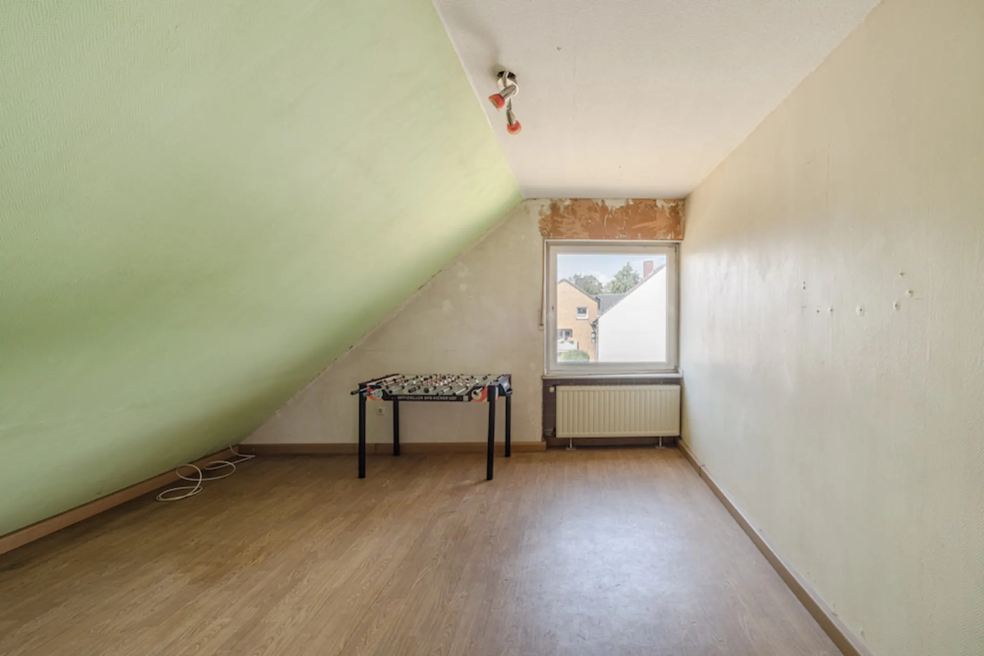 Dachzimmer mit schräger Decke, Fenster, Heizkörper und Kickertisch auf Laminatboden.