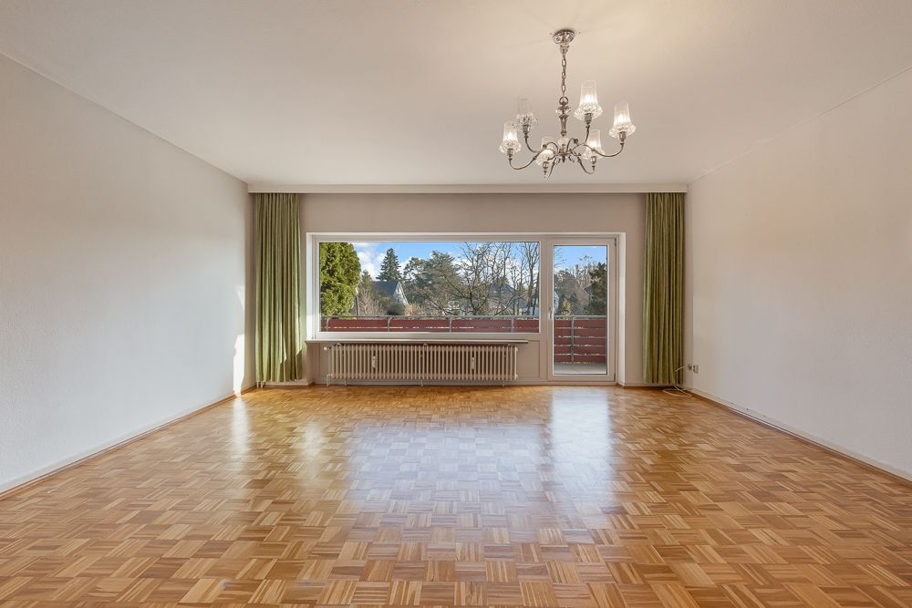 Leeres Wohnzimmer mit Parkettboden, großem Fenster, Tür zum Balkon und Kronleuchter.
