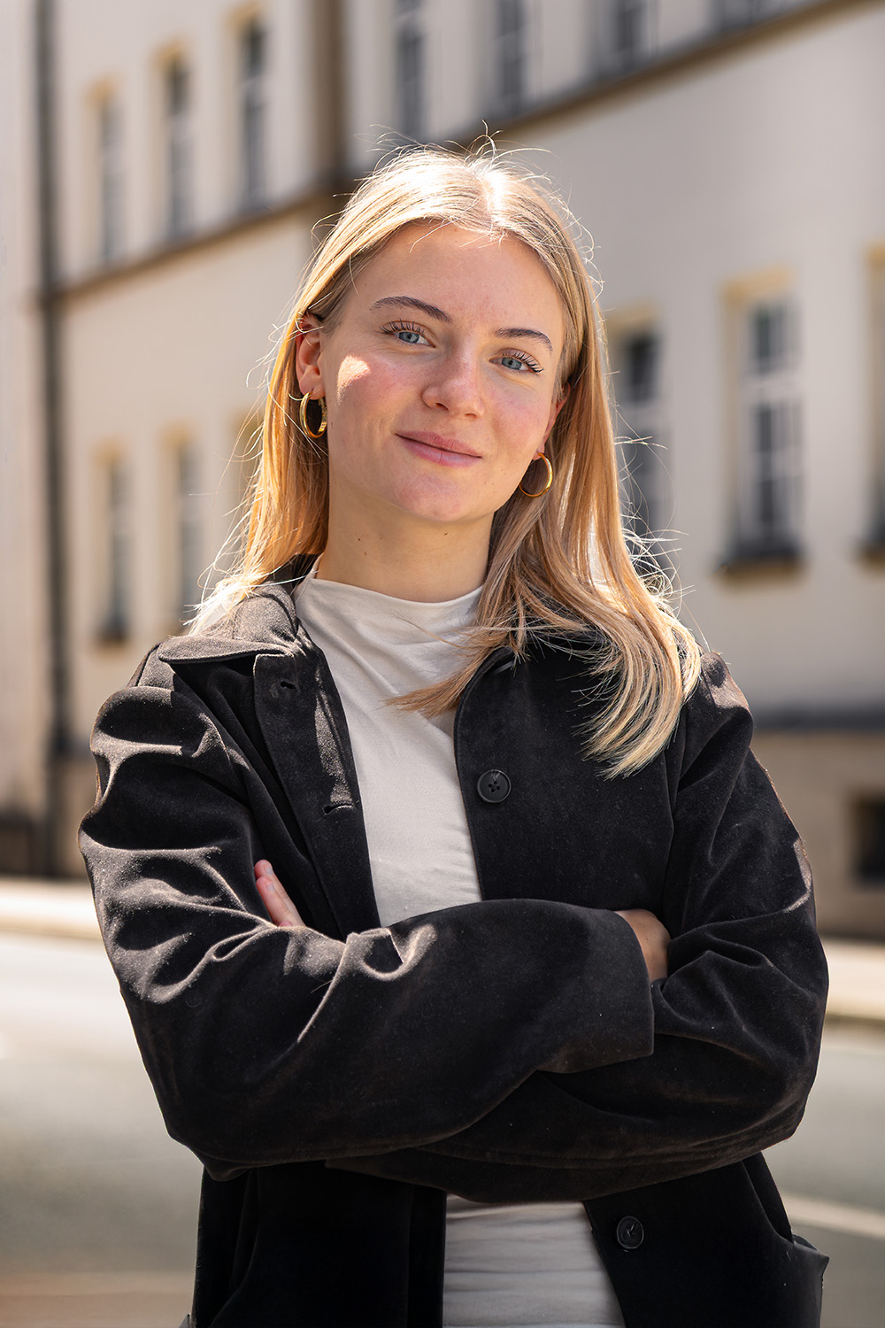 Junge Frau mit langen blonden Haaren steht mit verschränkten Armen vor einem Wohngebäude.
