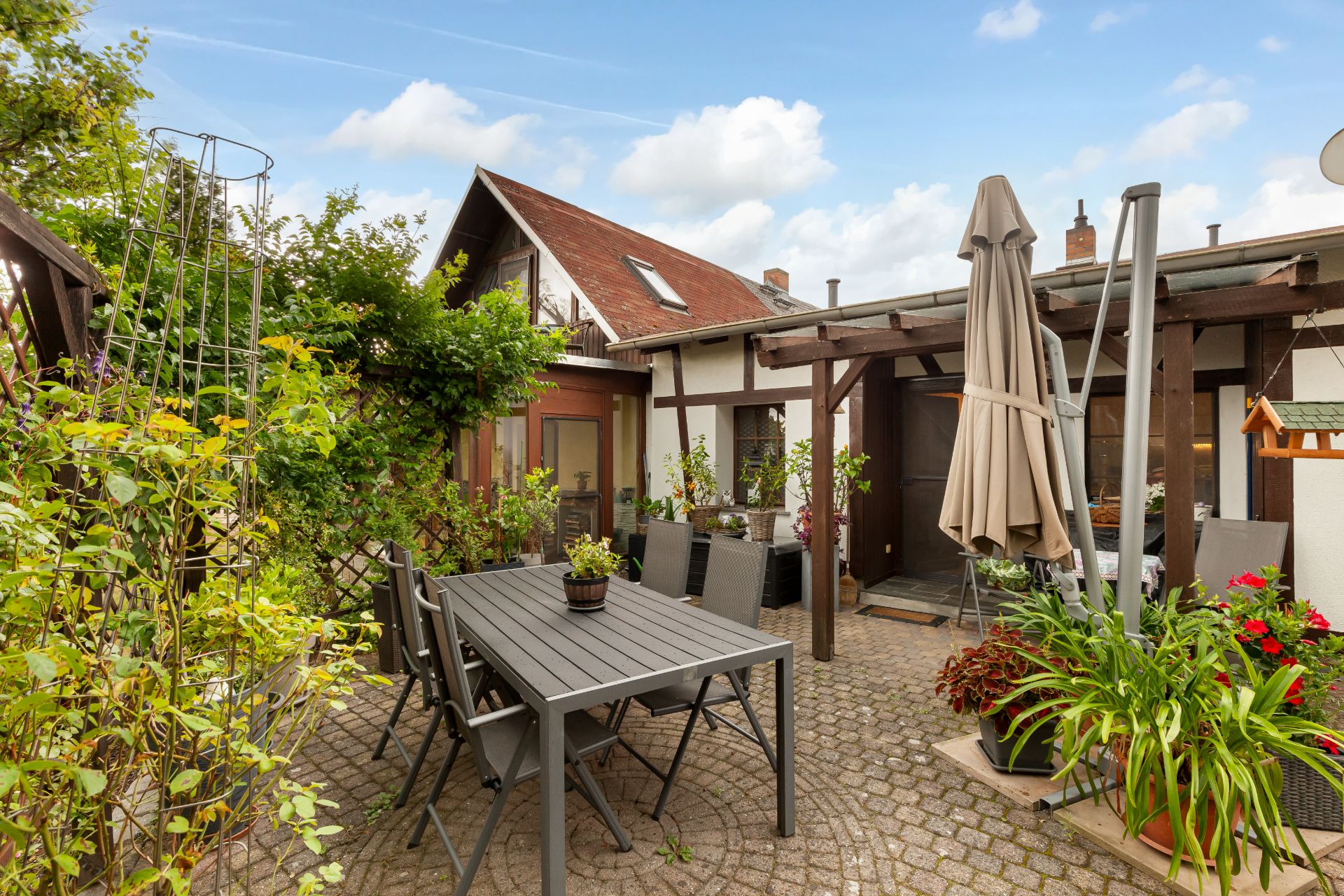 Gepflasterter Innenhof mit Gartentisch, Stühlen, vielen Pflanzen und Sonnenschirm vor einem Wohnhaus.