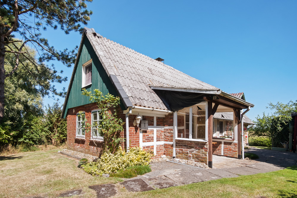 Backsteinhaus mit schrägem Dach, grünem Giebel und kleinem Garten an einem sonnigen Tag.