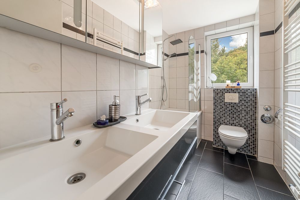 Modernes Badezimmer mit Doppelwaschbecken, WC, großer Dusche und Fenster mit Blick ins Grüne.