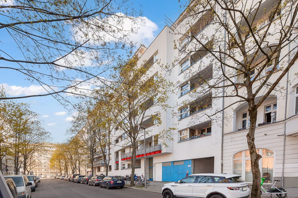 Mehrstöckiges Wohnhaus mit Balkonen an einer von Bäumen gesäumten Straße, geparkte Autos am Straßenrand.