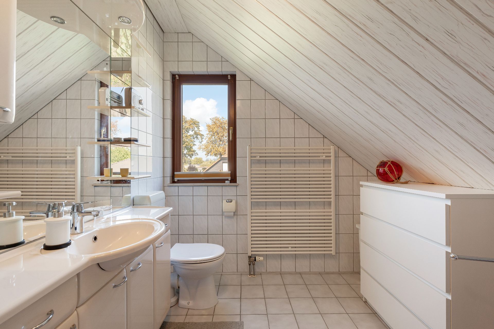 Helles Bad mit Dachschräge, Waschbecken, WC, Fenster, Handtuchheizkörper und weißer Kommode.