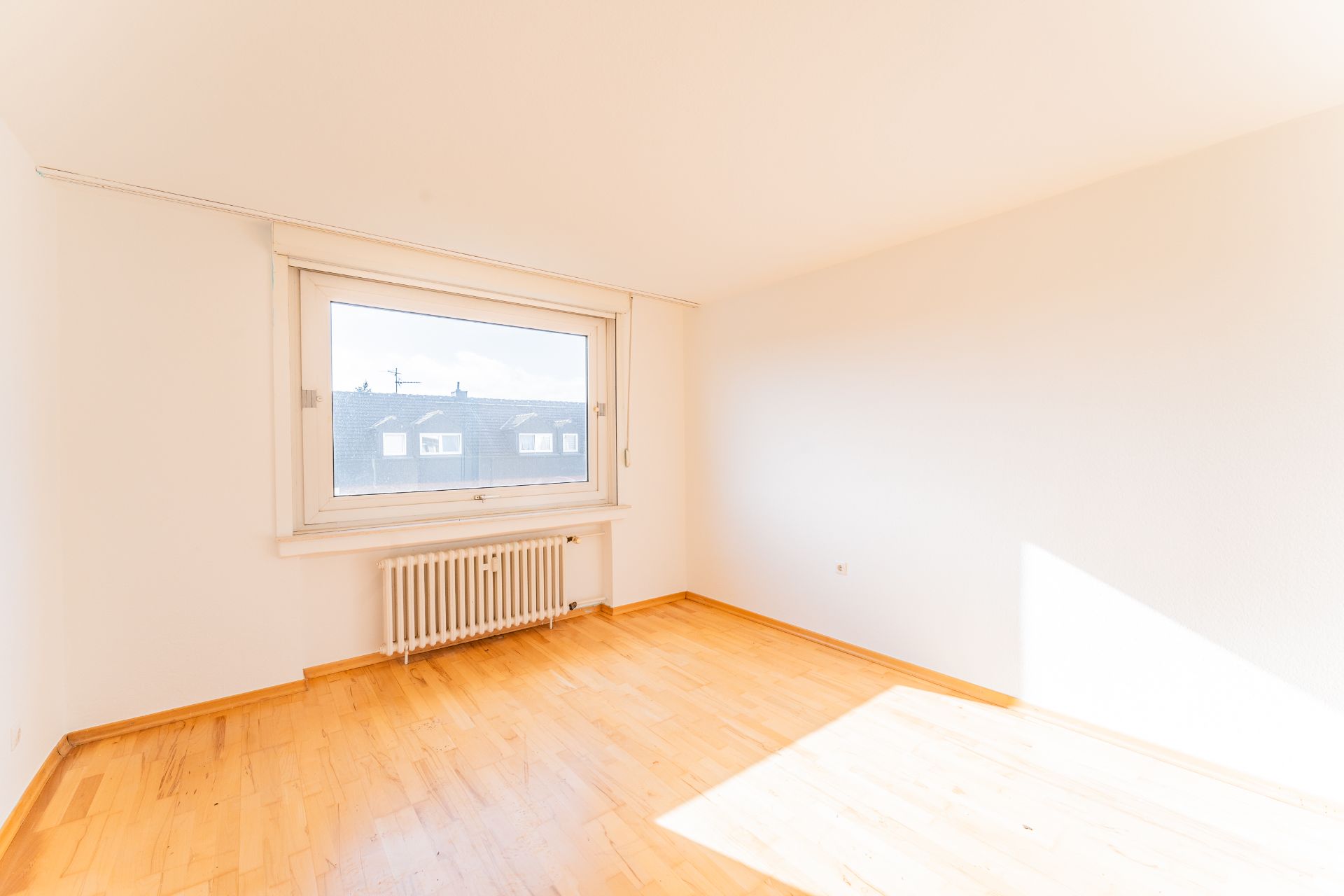 Helles, leeres Zimmer mit großem Fenster, Heizkörper und hellem Holzboden.