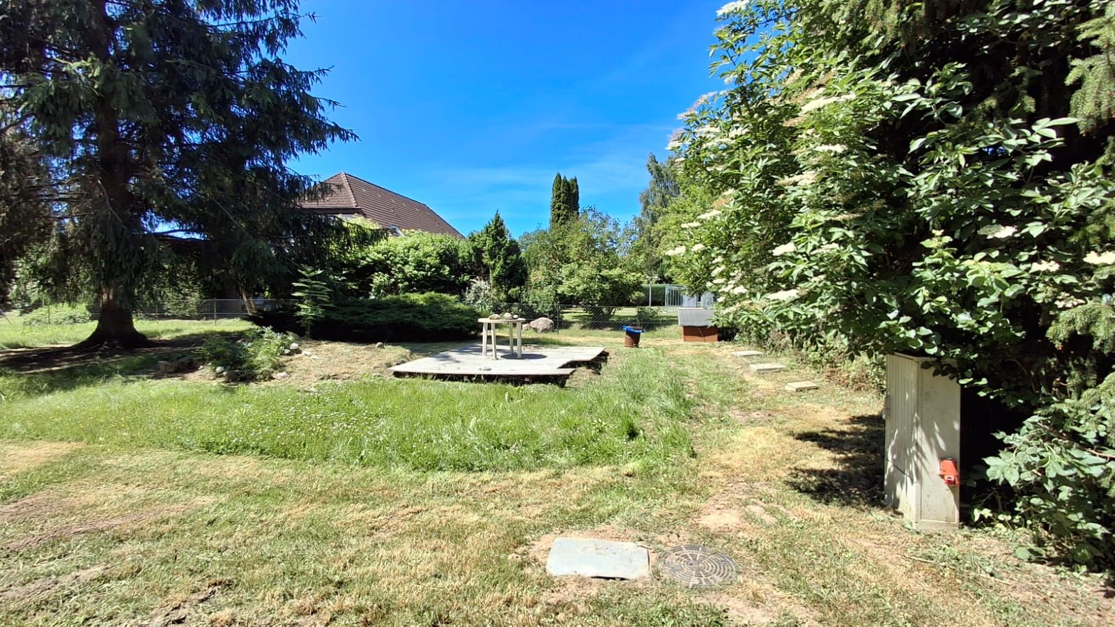 Garten mit Holzterrasse, Rasen, Bäumen und Sträuchern bei sonnigem Wetter.