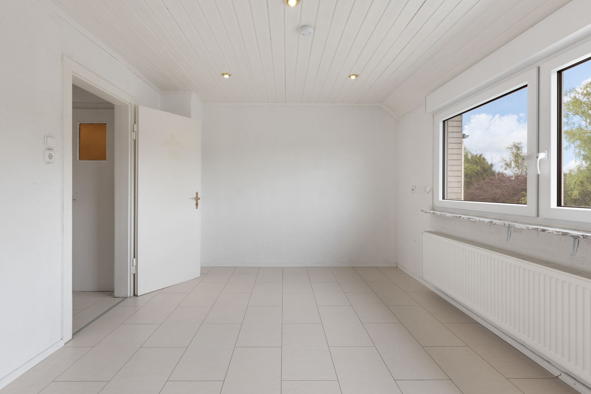 Heller, leerer Raum mit weißen Wänden, Fliesenboden und großem Fenster.
