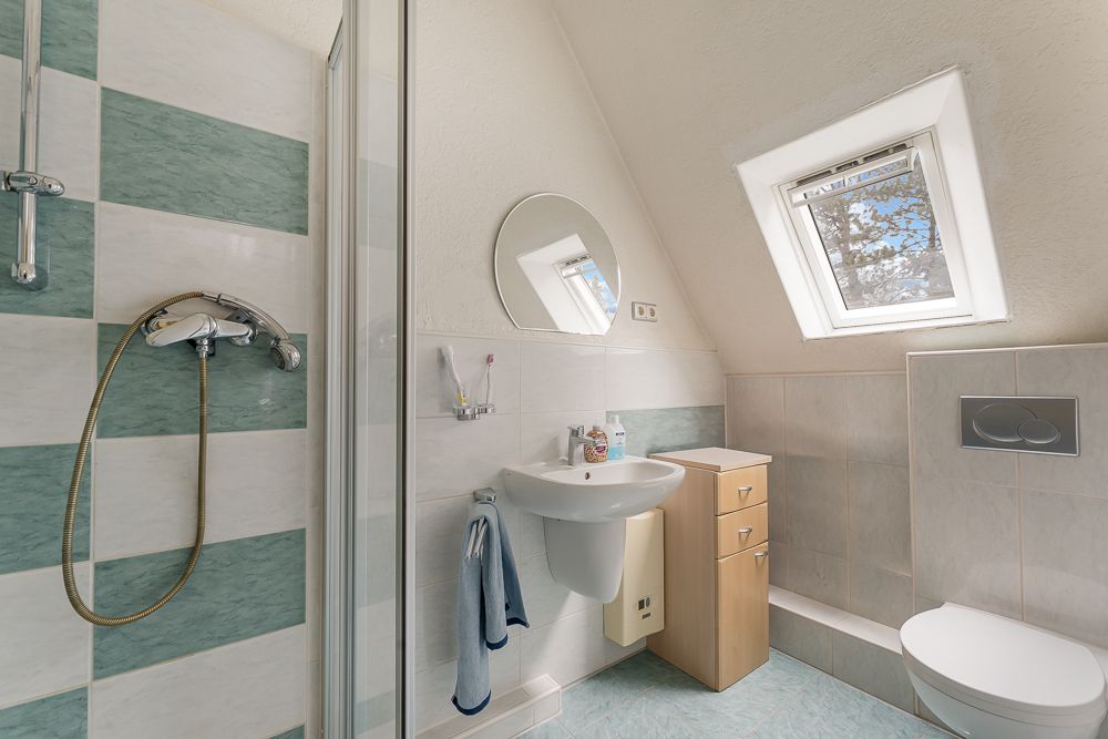 Modernes Bad mit Dusche, Waschbecken, Spiegel, Fenster, Schrank und WC unter schrägem Dach.