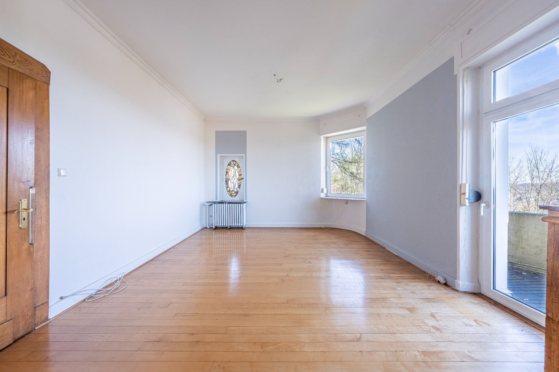 Leeres Wohnzimmer mit Holzboden, großen Fenstern und Holztür, Zugang zu einem Balkon.