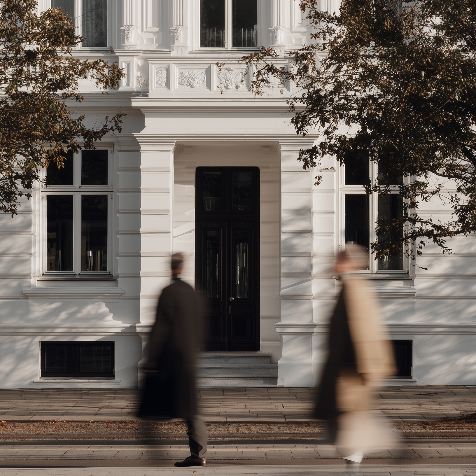 Fassade eines weißen Wohngebäudes mit verzierten Fenstern und vorbeigehenden Personen.