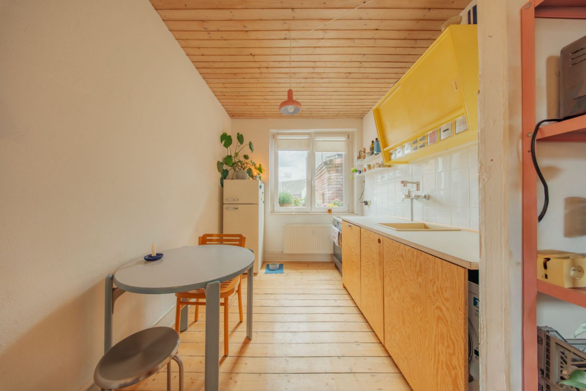 Helle Küche mit Holzboden, gelben Schränken, Esstisch und zwei Stühlen, Fenster im Hintergrund.