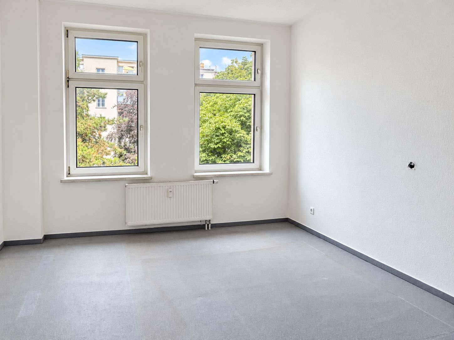 Helles, leeres Zimmer mit drei Fenstern und Heizkörper an der Wand.