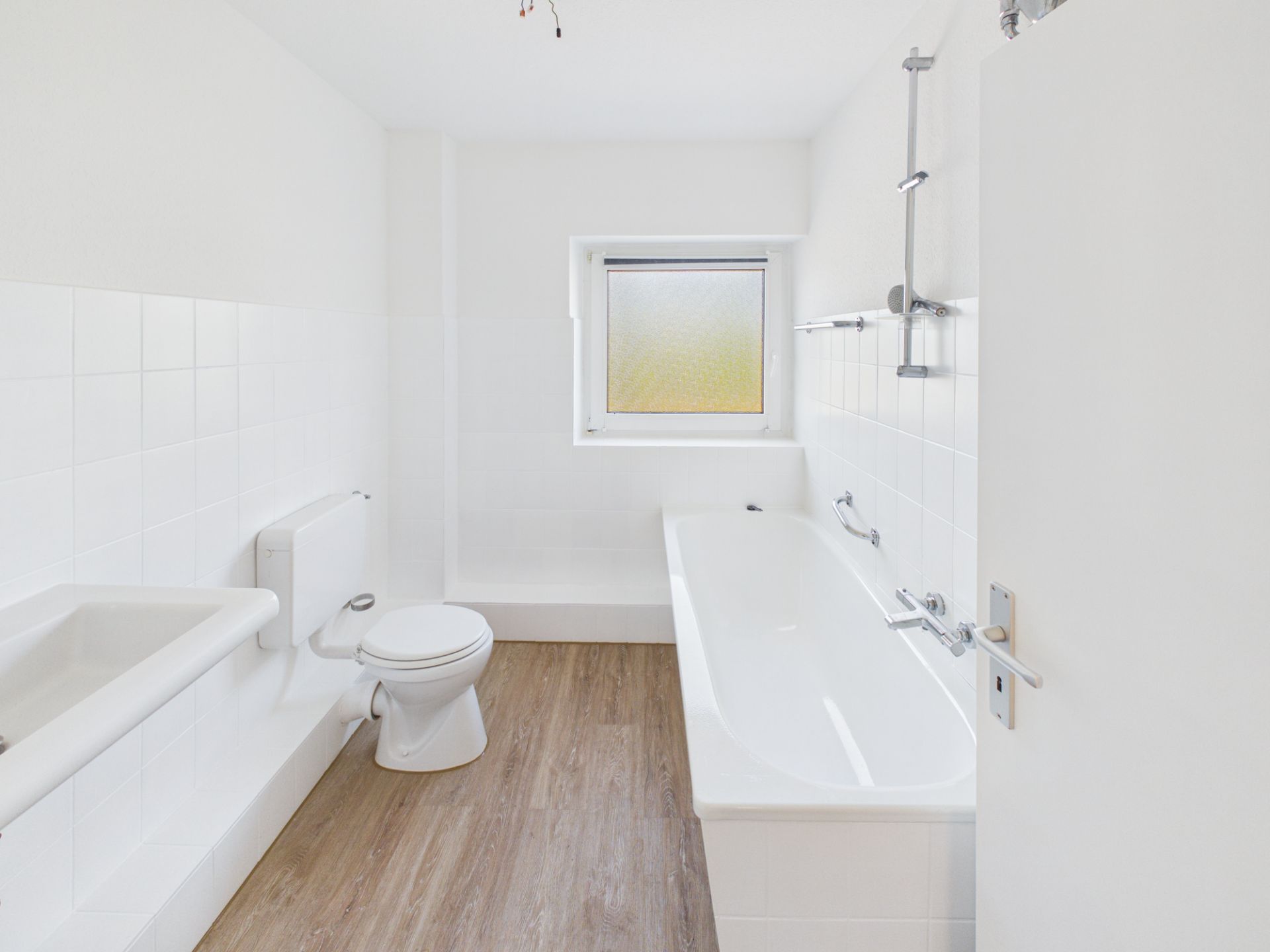 Helles Badezimmer mit Badewanne, Toilette, Waschbecken und Fenster, Boden in Holzoptik.