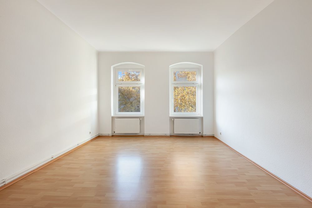 Leerer, heller Raum mit Holzfußboden und zwei Fenstern mit Heizkörpern darunter.